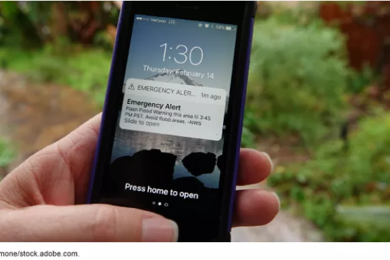 A hand holding a cell phone displaying an emergency weather alert from the National Weather Service 