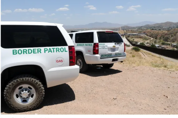 Two white SUVs parked in the dirt next to a road, with a long fence on the right-hand side of the road.