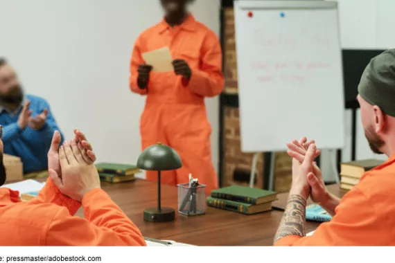 One person in an orange jumpsuit stands next to a whiteboard on an easel, while two people in orange jumpsuits and one person in a blue shirt sit around a table and applaud.