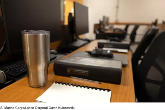 A row of empty desks and chairs with an unattended cup of coffee and binders in the foreground, with additional empty desks in the background.
