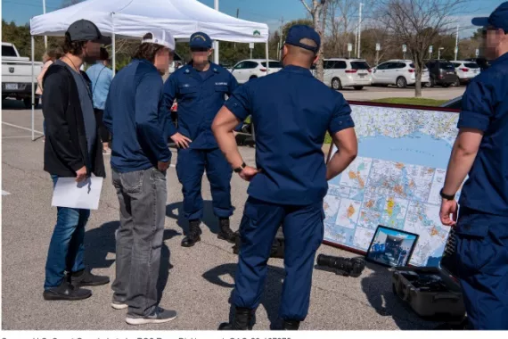 A group of people gathered on an outside parking lot on a sunny day. Coast Guard personnel in dark blue uniforms are talking to people. A large map is propped up on the ground. 