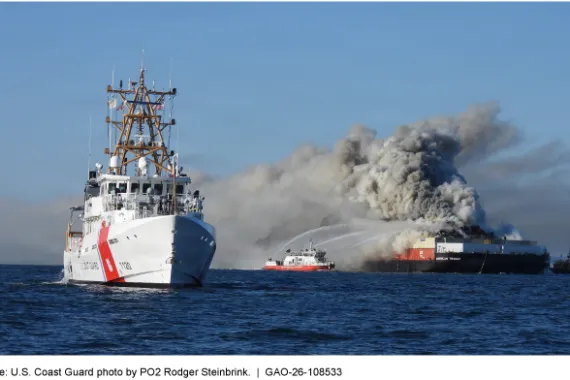 A large white ship in the foreground with a smaller boat in the background spraying water on a larger ship with smoke billowing off it.