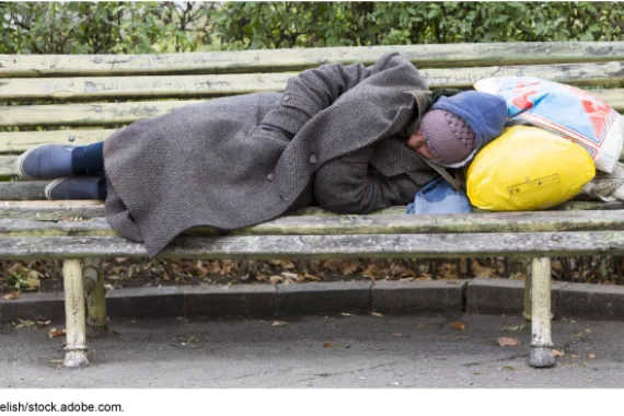 A person sleeping on a bench in a long trench coat and winter hats with bags as a pillow.