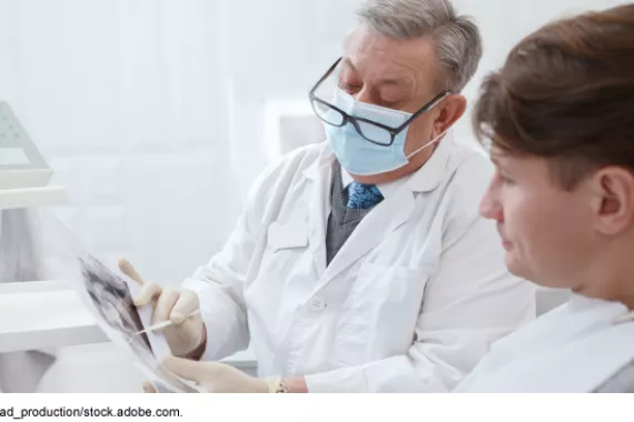 A person wearing glasses and a doctor’s white coat holding papers sitting next to another person sitting in a chair in a dental office setting.