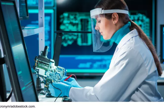 A person in a white coat and a clear face shield works in a laboratory on a circuit board.