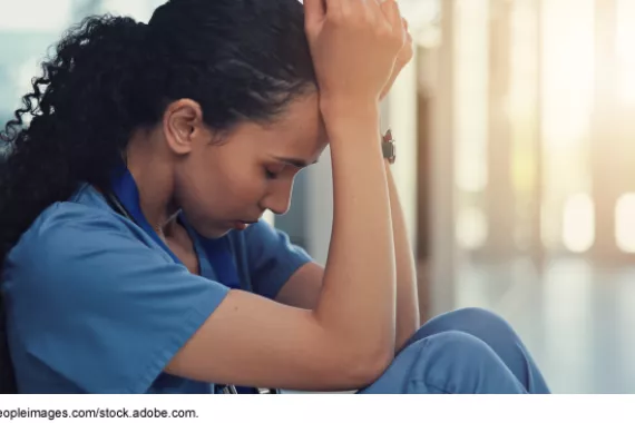 A woman with curly hair, wearing short-sleeved hospital scrubs, sits on the floor with her head in her hands.