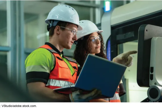 Two people in reflective vests, hard hats, and safety glasses are looking at computers. 