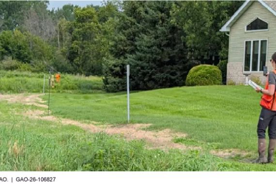 A U.S. Army Corps of Engineers official standing on a pathway in grass next to a home, holding up a handheld device.