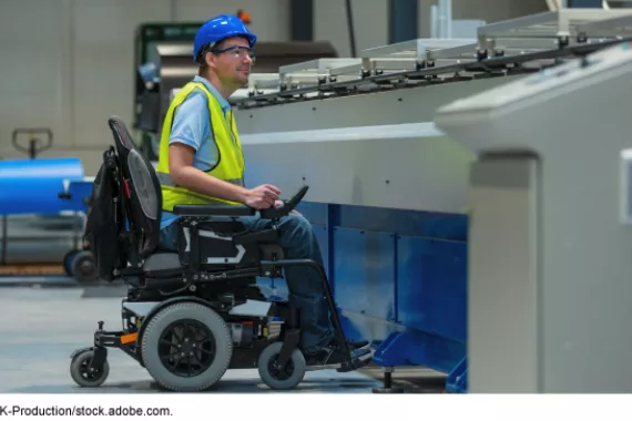 A man in an electric wheelchair, wearing safety glasses, a yellow vest and blue helmet in a factory.