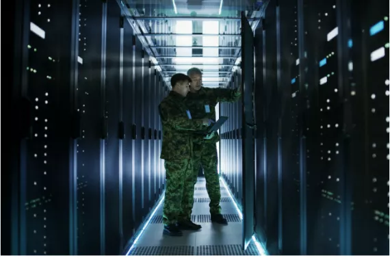 In a data center, two military men work with an open server rack cabinet. One holds a military edition laptop.