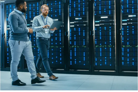 Two men, one carrying a laptop, walking in a server room