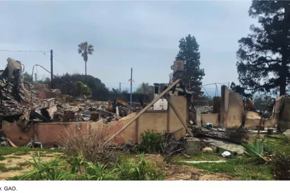 Partial burnt remains of a house’s walls and frames in the aftermath of a fire. Large piles of rubble are in the background. 
