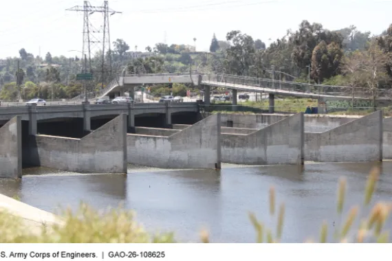 A bridge with cars on it over a river with several large concrete structures running parallel in the river.