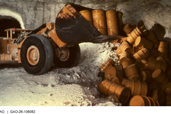 Inside an underground chamber with rock walls, a large industrial vehicle moves several yellow cylindrical drums that resemble barrels to a pit that is partially filled with other drums.