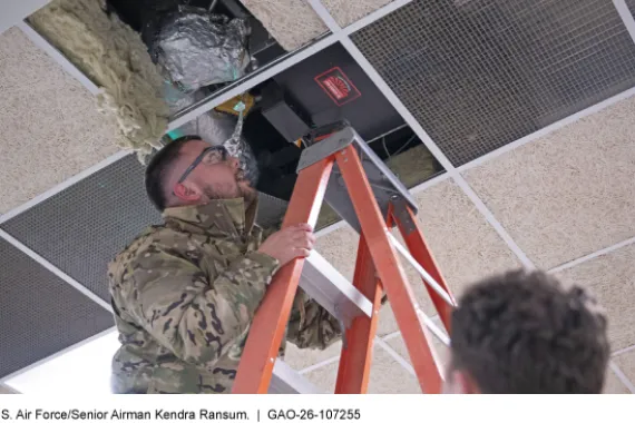Airman on a ladder inspects equipment above ceiling tiles.
