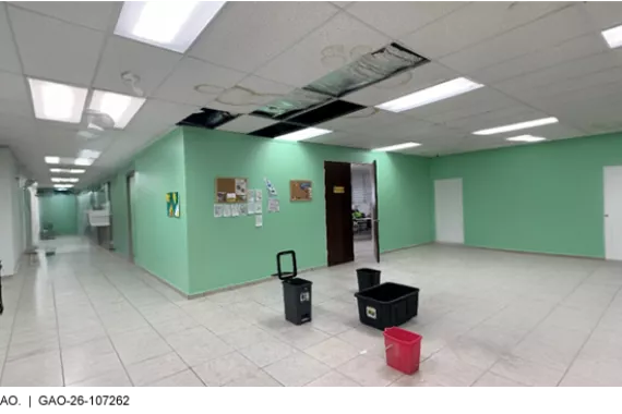 An open space with tile flooring and visible water damage on the drop ceiling above. Some ceiling tiles are missing, and the floor has a few plastic bins on it to catch water.