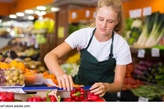 Teenage grocery worker wearing an apron, near boxes of red peppers.