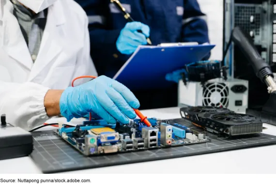 A person in a white coat and blue latex gloves is pointing to something on a computer motherboard with a tool while a person in the background holds a pen to a clipboard.