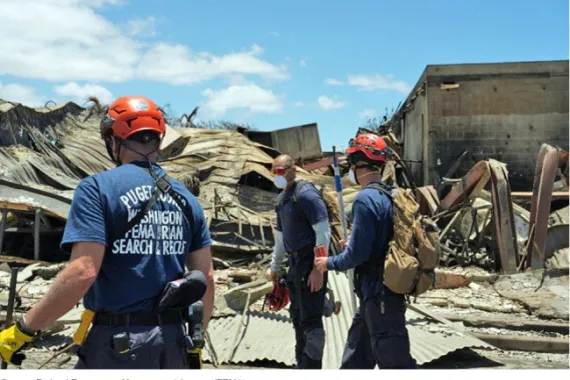 Three men wearing or carrying hard hats, masks, gloves, packs, and utility belts stand near the remains of a destroyed building.