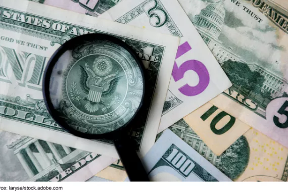 A pile of U.S. paper money of different denominations with a magnifying glass over the great seal on the back of the $1.