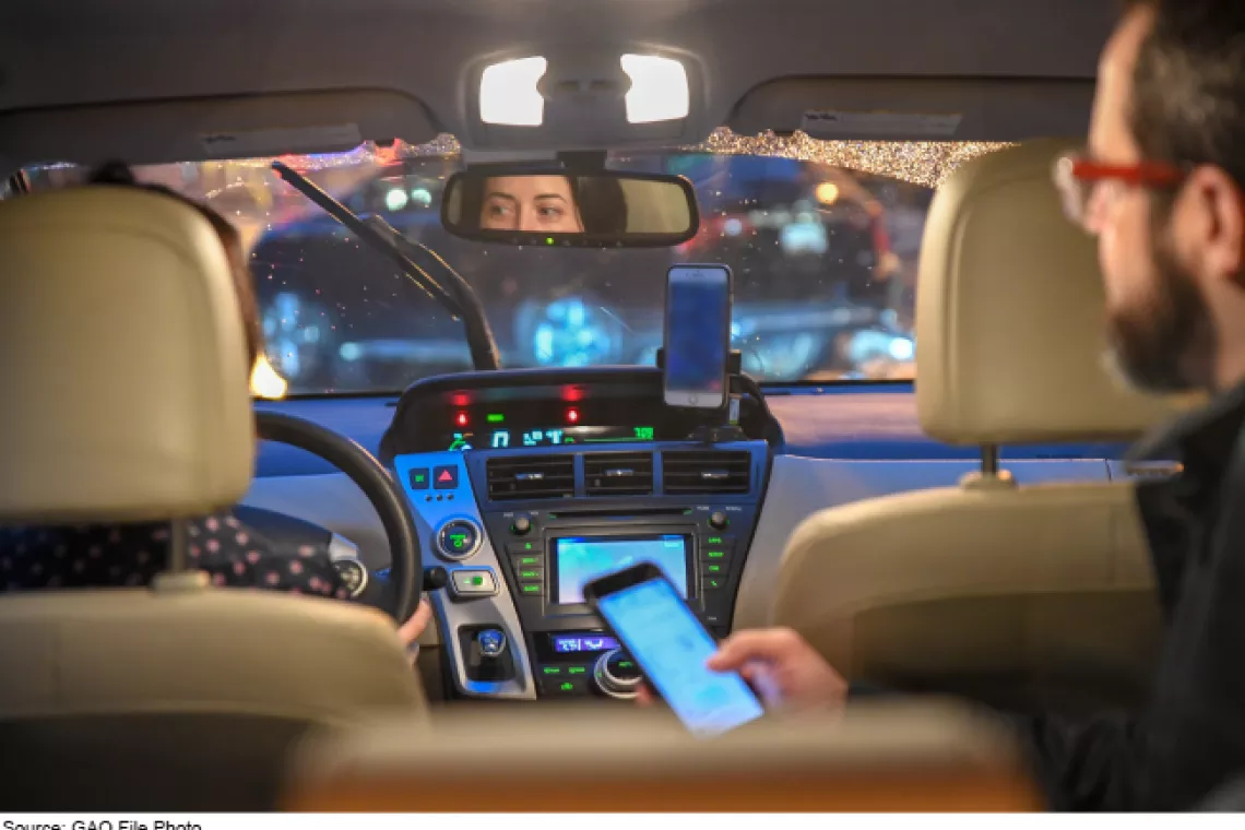 Image showing the inside of a car with one person behind the wheel and another in the backseat holding a phone.