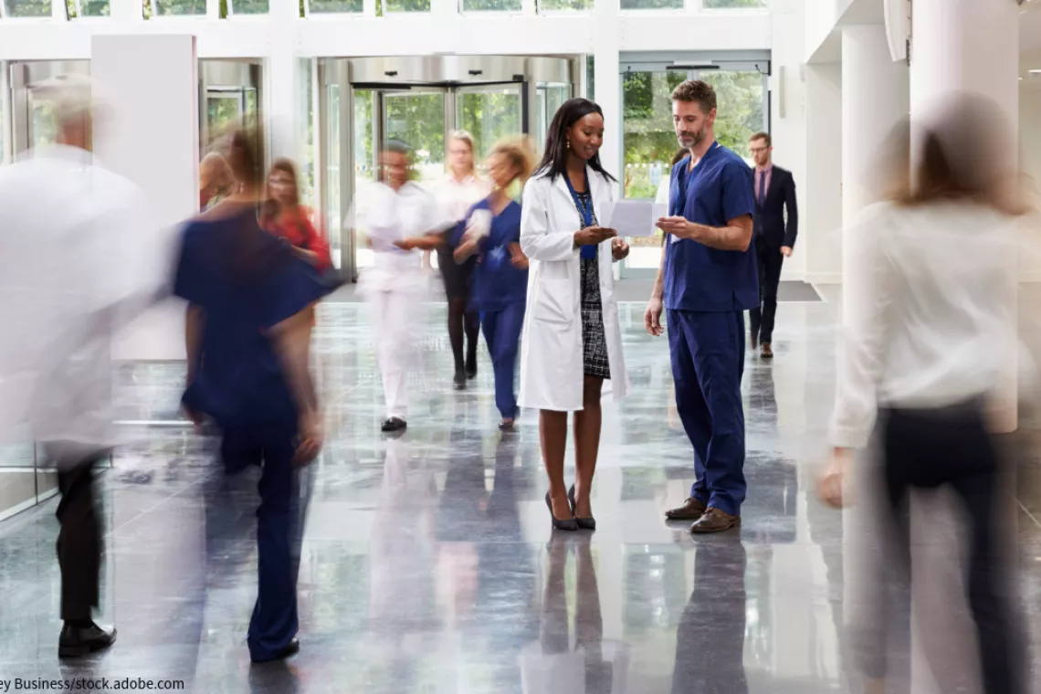 Image shows two doctors having a discussion in the midst of a busy hospital hallway as other doctors, nurses, and healthcare professionals walk past